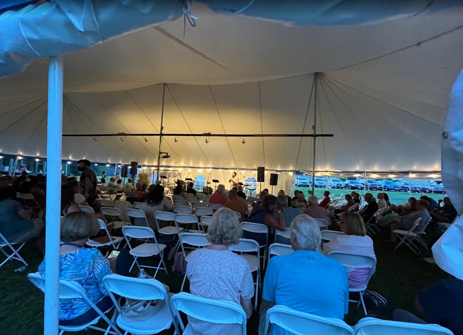 Guests seated in white folding chairs under a lit party tent, enjoying an evening gathering