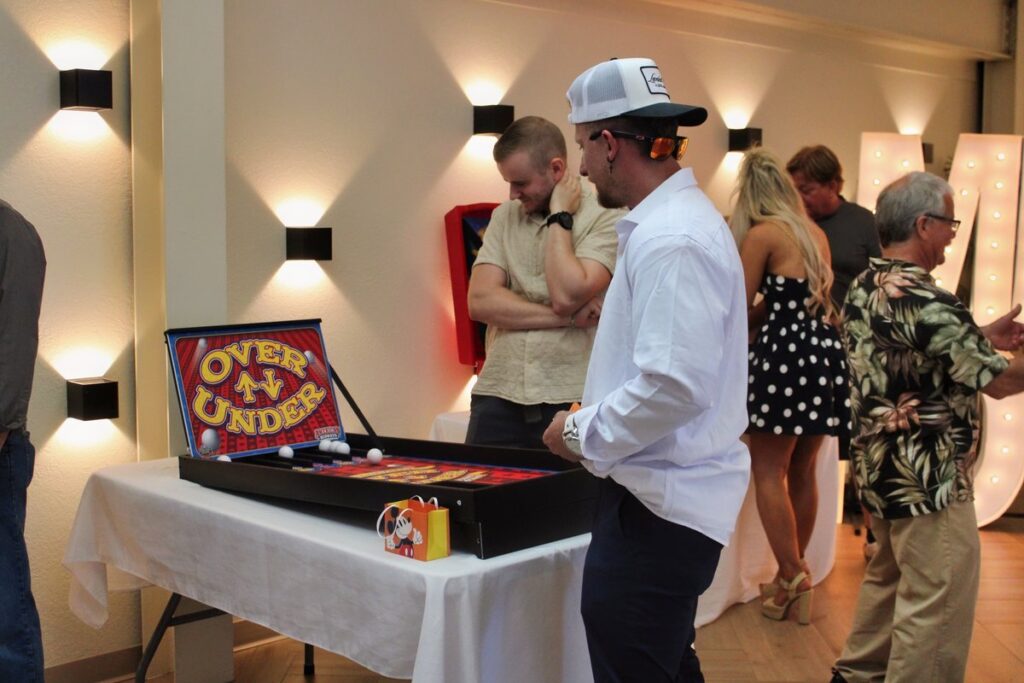 Guests of all ages gathered around an Over Under game at an indoor holiday party with a white linen table, from Just In Case Party Rentals